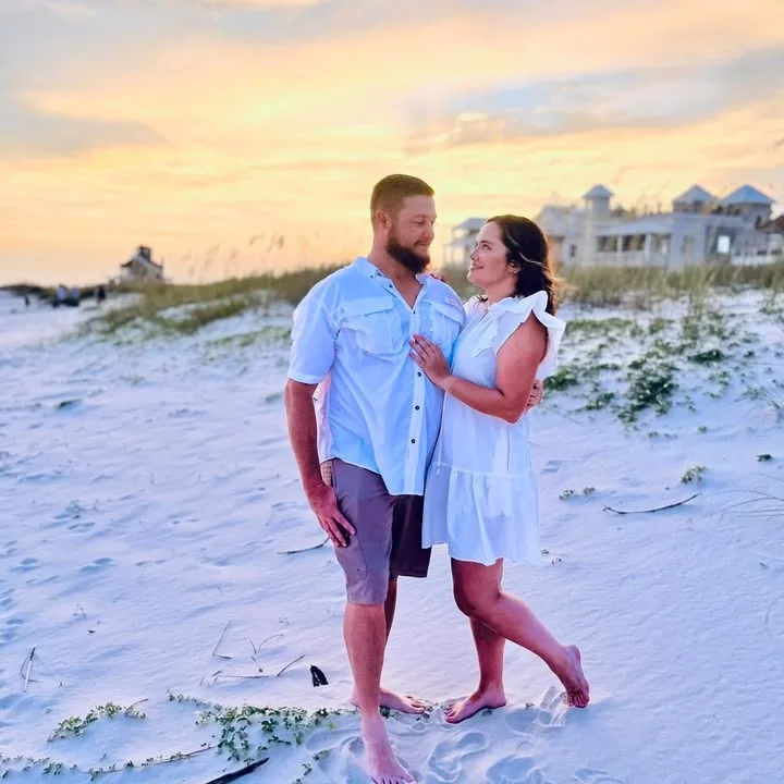 Danny and Diedre standing together on a beach at sunset, smiling at each other