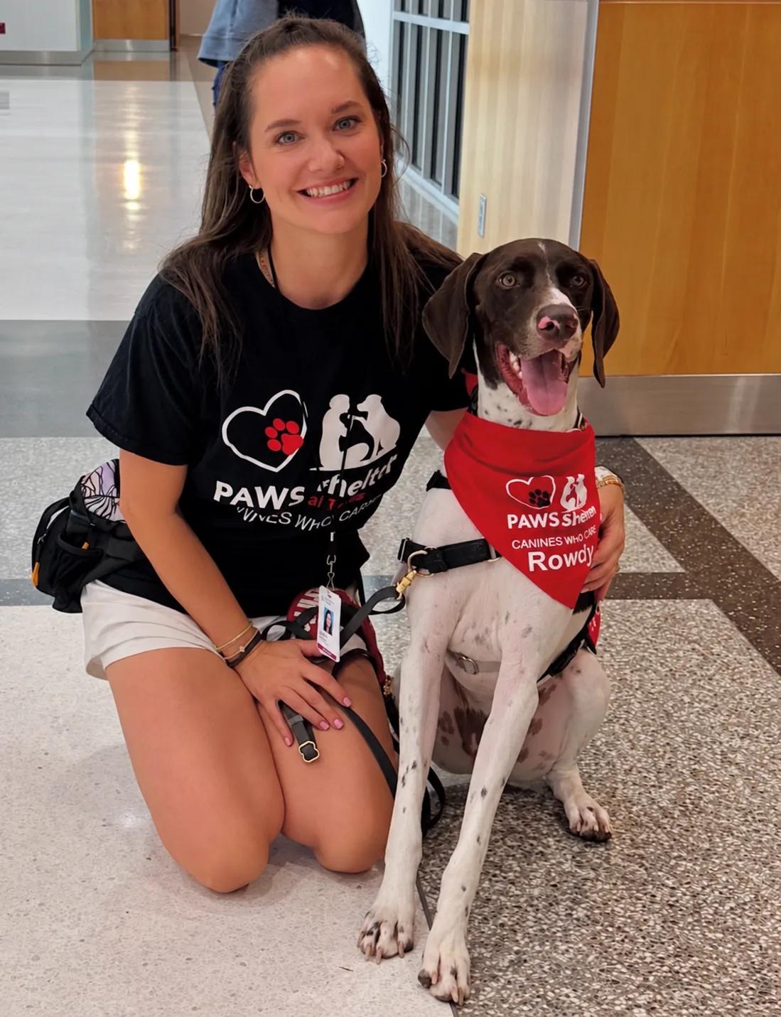 Diedre kneeling beside a smiling Rowdy in a hallway, with his red Canines Who Care bandana